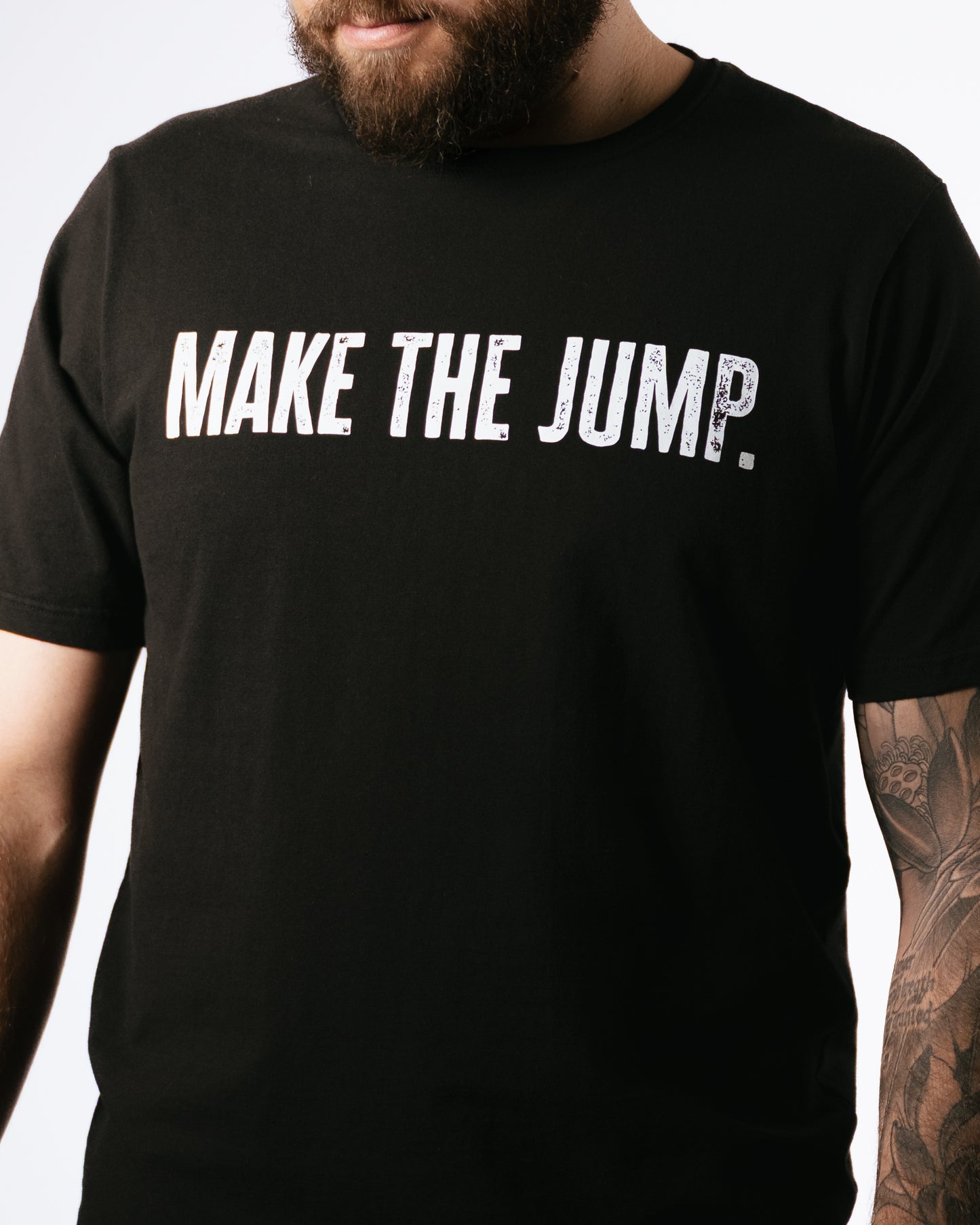 Black t-shirt with 'MAKE THE JUMP.' text worn by a person with tattoos on a white background